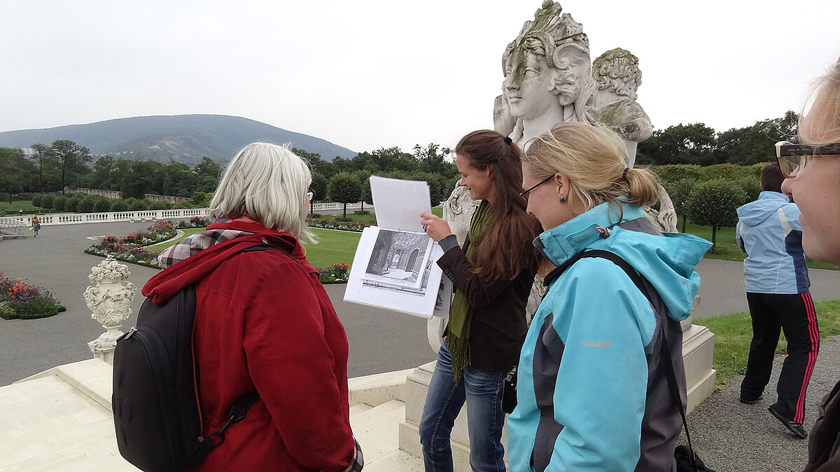 Drei Frauen stehen bei einer Sphinx Statue beim Schloss Hof. Eine der Frauen zeigt eine historische Aufnahme.