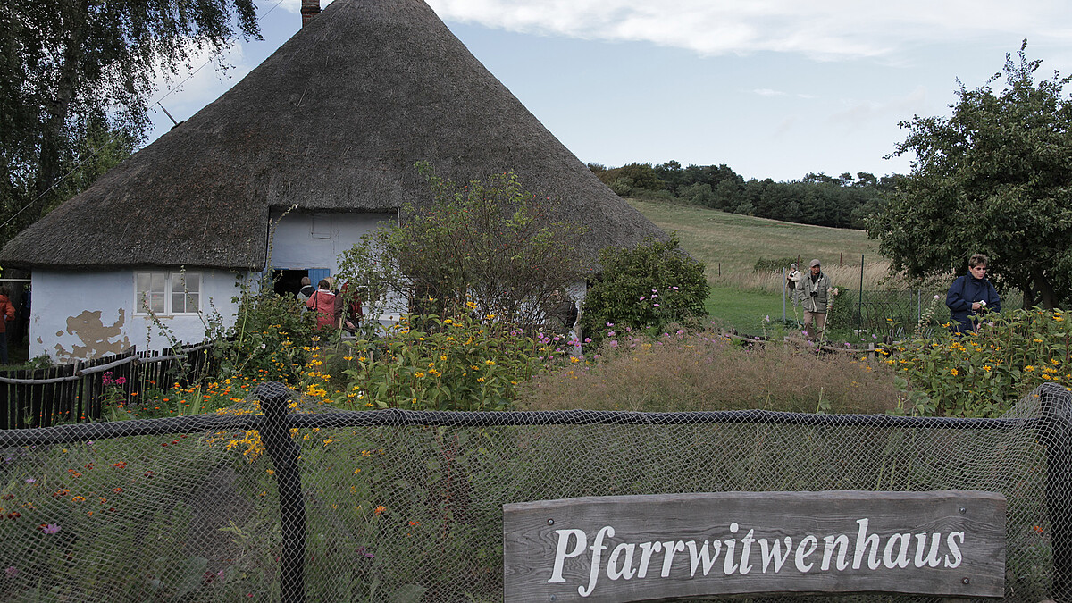 Ein paar Personen besichtigen ein weißes Haus mit Reetdach und Garten. Am Gartenzaun ist ein Schild mit der Aufschrift "Pfarrwitwenhaus" angebracht.